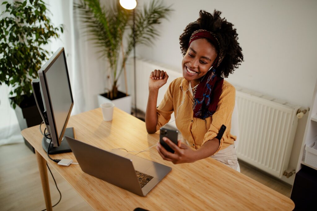 Young black woman answering a call on Internet-enabled smart phone while working on laptop.